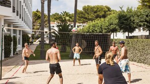 A group of people playing beach volleyball on a sandy court with a net, surrounded by greenery and buildings.
