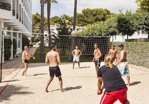 Un grupo de personas jugando voleibol de playa en una cancha de arena, rodeado de vegetación y edificios.