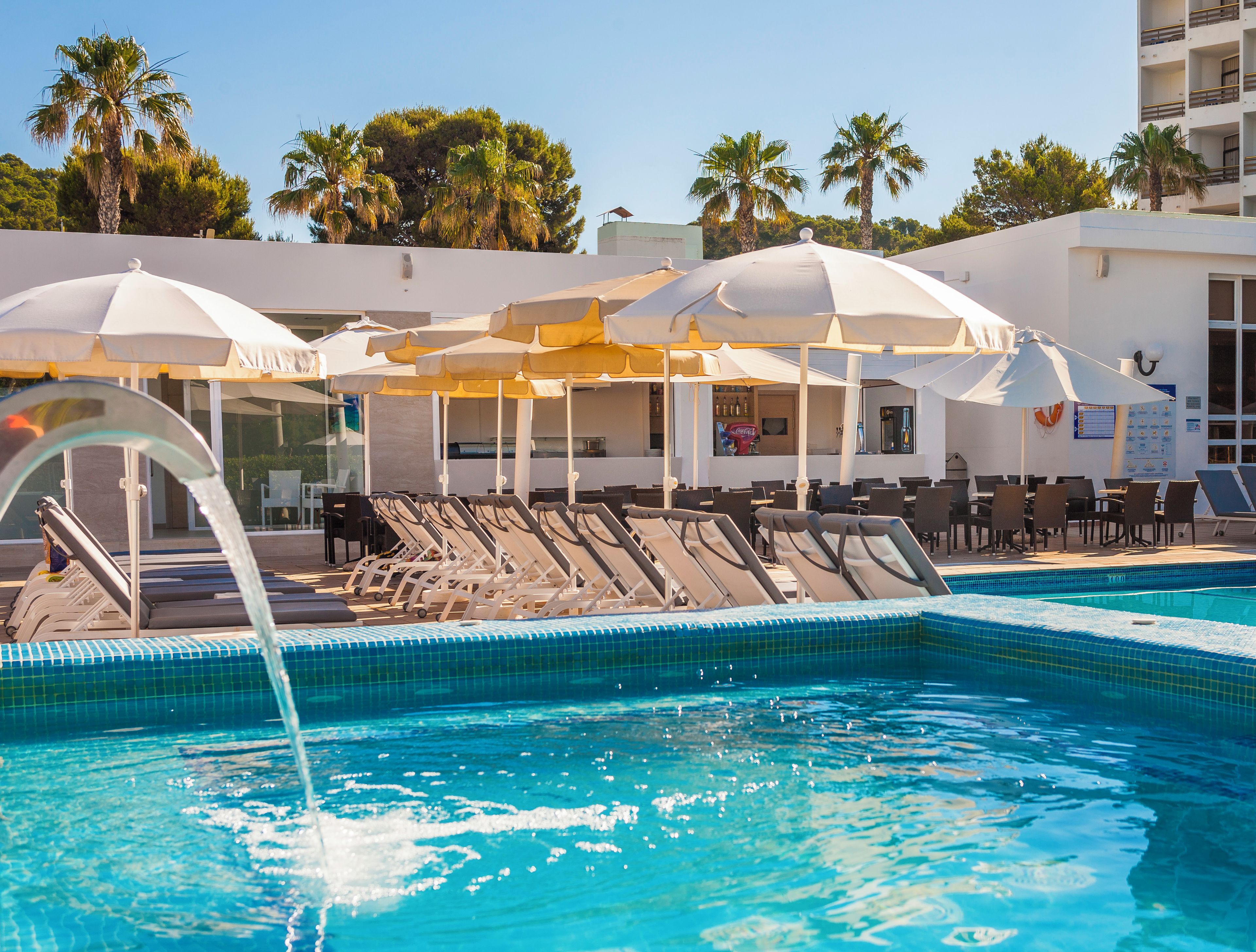 A pool area with lounge chairs and umbrellas, a bar, and a view of palm trees and a building.