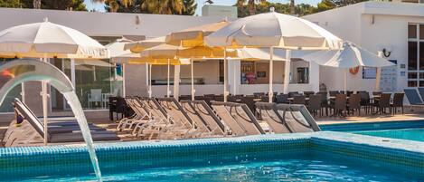 A pool area with lounge chairs and umbrellas, a bar, and a view of palm trees and a building.