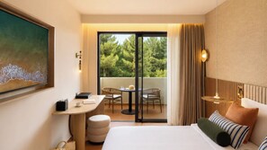 A hotel room with a bed, a desk, a chair, a small table, a sofa, a painting, and a view of trees through a sliding door. - Mitsis Selection Azure (Kassandra)