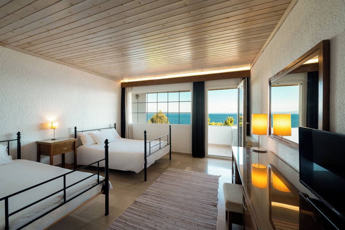 A hotel room with two beds, a TV, a desk, and a large window with a view of the sea.