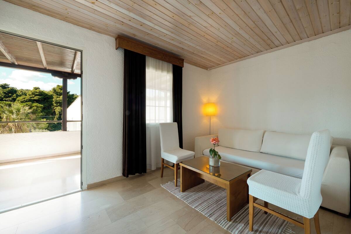 A modern living room with a white sofa, a wooden coffee table, and a chair. There is a window with curtains, a lamp on the wall, and a view of greenery outside.