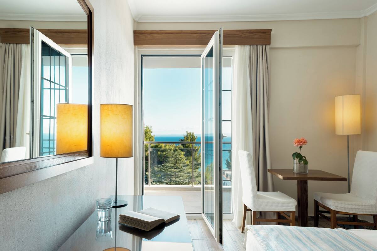 A hotel room with a balcony, a desk, two chairs, a bed, and a lamp.