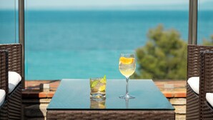 A table with two drinks and wicker chairs, overlooking a body of water. - Canvas by Mitsis Alexander the Great (Kassandra)