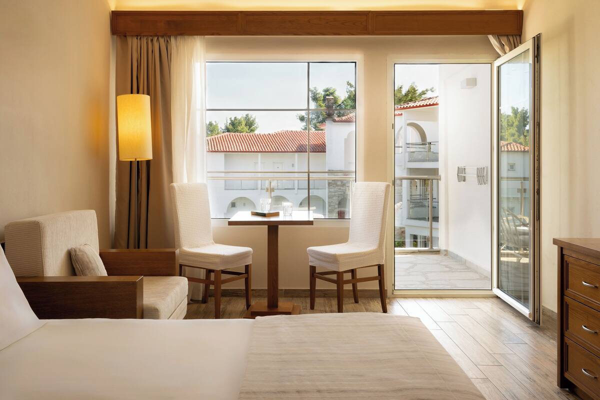A hotel room with a bed, two chairs, a table, a lamp, a balcony with a glass door, and a view of buildings.