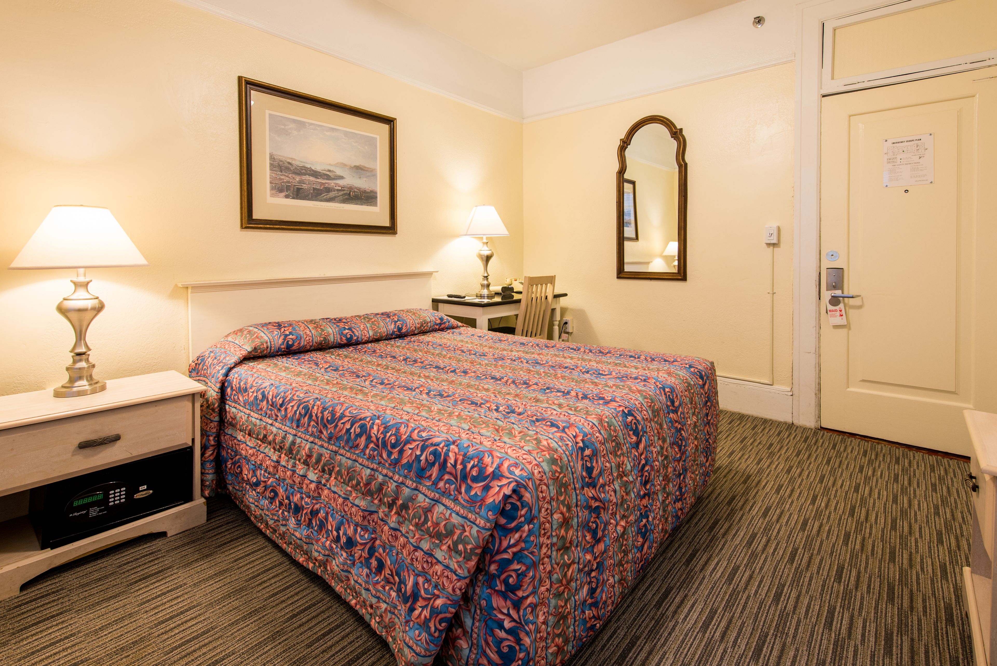 A hotel room with a bed, two bedside lamps, a desk with a chair, a framed picture on the wall, a mirror, and a door with a notice.
