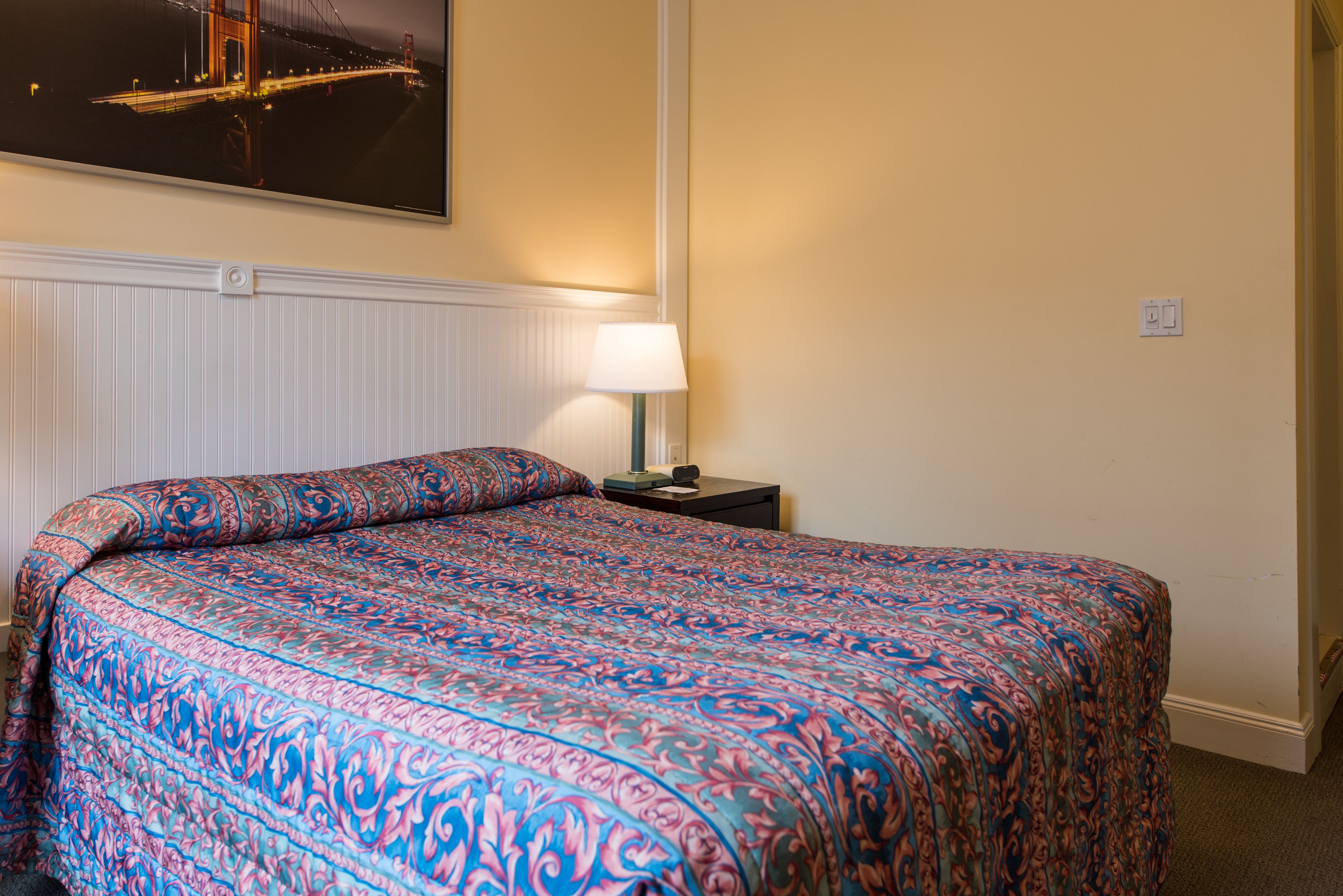 Una habitación de hotel con cama, mesita de noche, lámpara y televisor empotrado en la pared que muestra un paisaje urbano.
