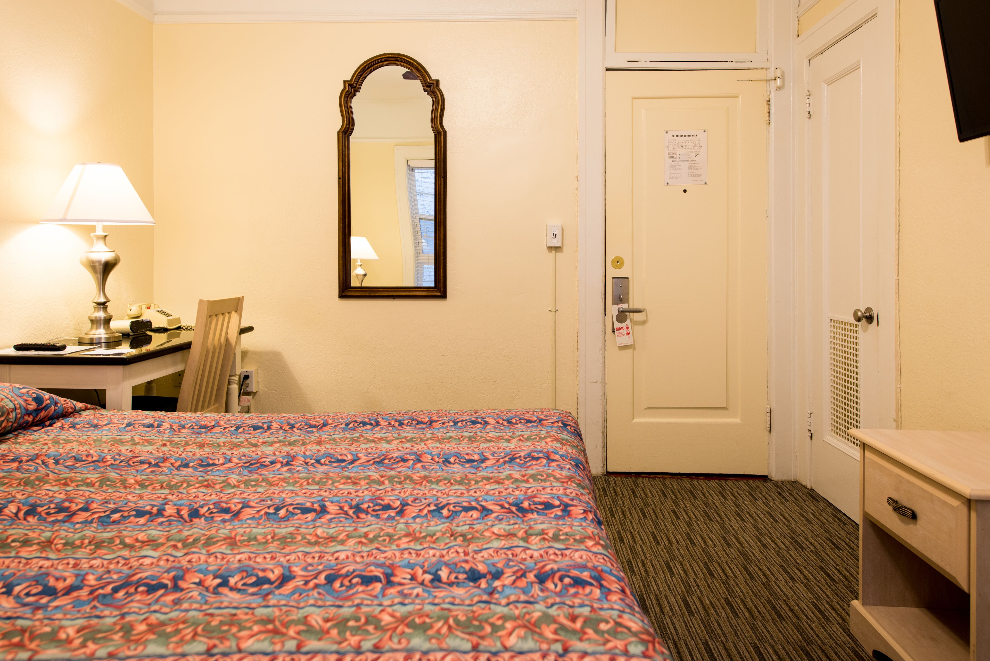A hotel room with a bed, a desk with a lamp, a chair, a mirror, and a door.