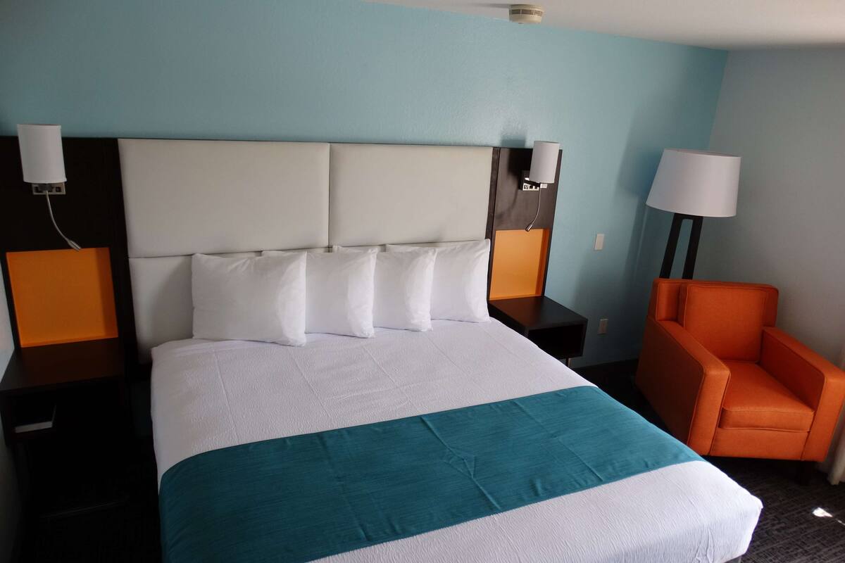 Ein Hotelzimmer mit einem großen Bett, zwei Nachttischen, einem orangefarbenen Sessel und einem Beistelltischchen mit einer Lampe.