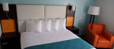 A hotel room with a large bed, two bedside tables, an orange armchair, and a nightstand with a lamp.