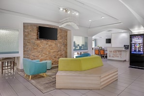 A modern hotel lobby with a stone wall, a flat-screen TV, a yellow sofa, teal armchairs, a reception desk, and a digital information kiosk.