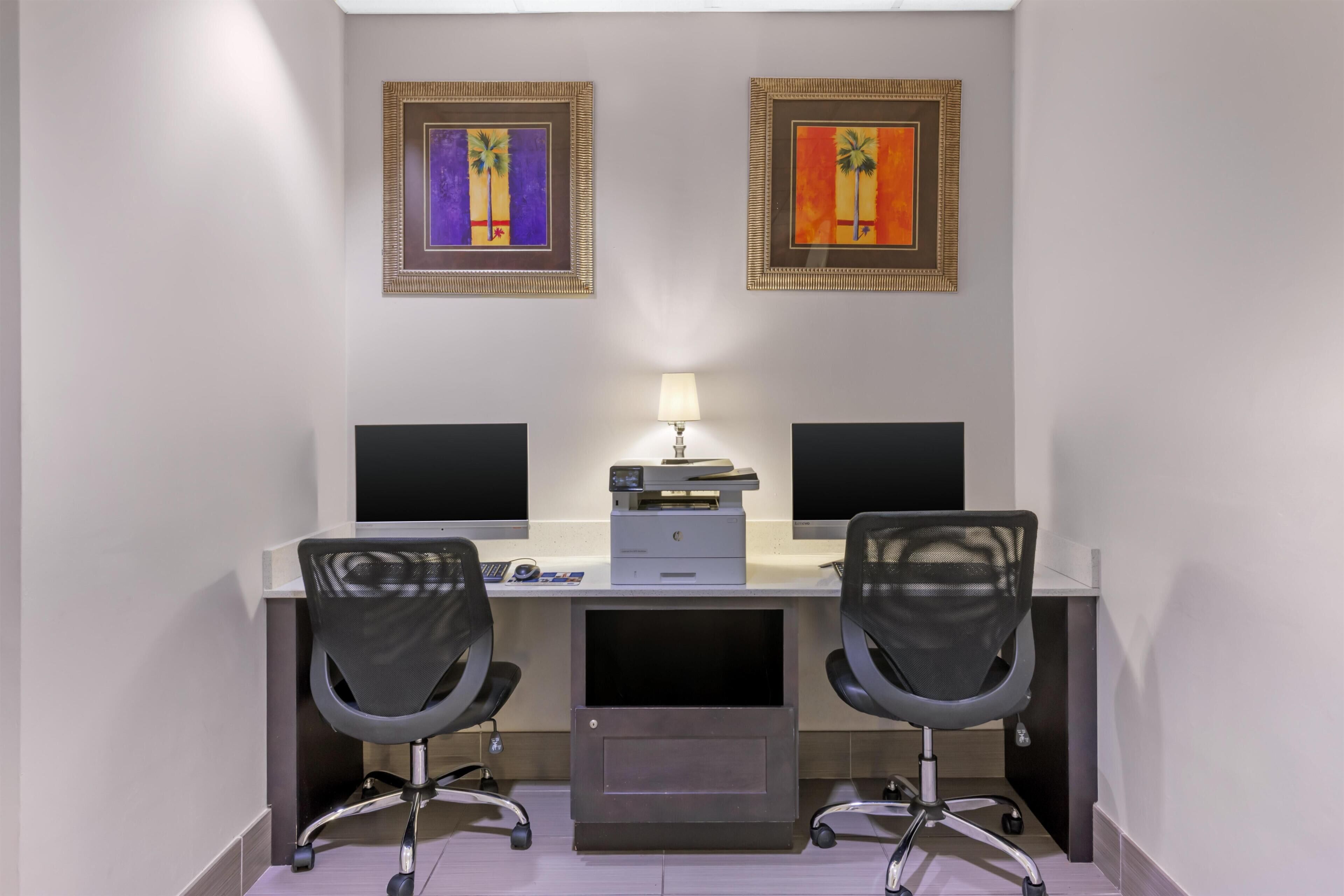 A room with two desks, two chairs, two monitors, a printer, and two framed artworks on the wall.