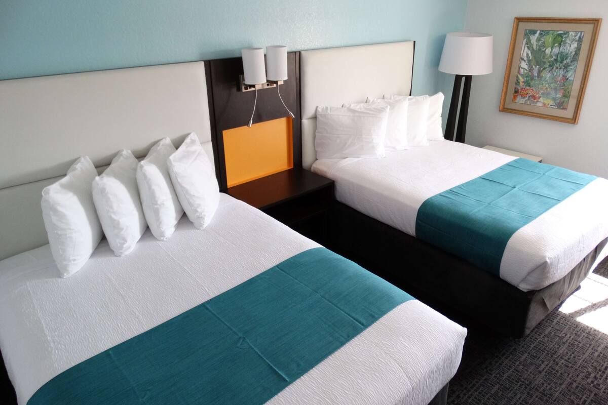 Ein Hotelzimmer mit zwei Betten, jeweils mit weißer Bettwäsche und Türkis-Akzenten, einem Nachttisch mit Lampe sowie einem Bild an der Wand.