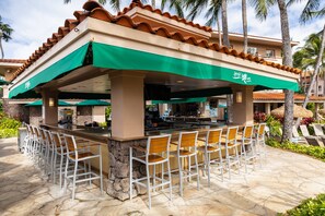 An outdoor bar area with a stone floor, white metal chairs, and a green awning.