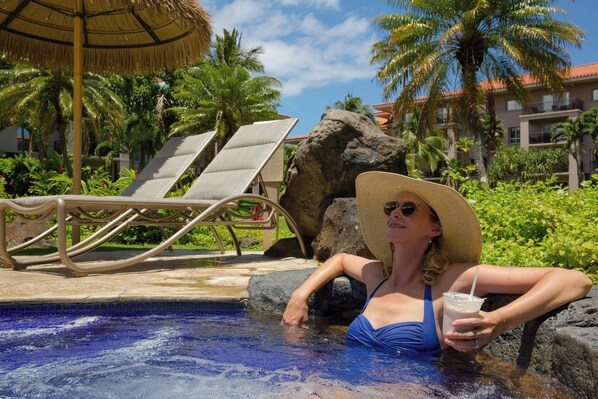 Un área de piscina con sillones de descanso, un entorno tropical con palmeras y un edificio al fondo.