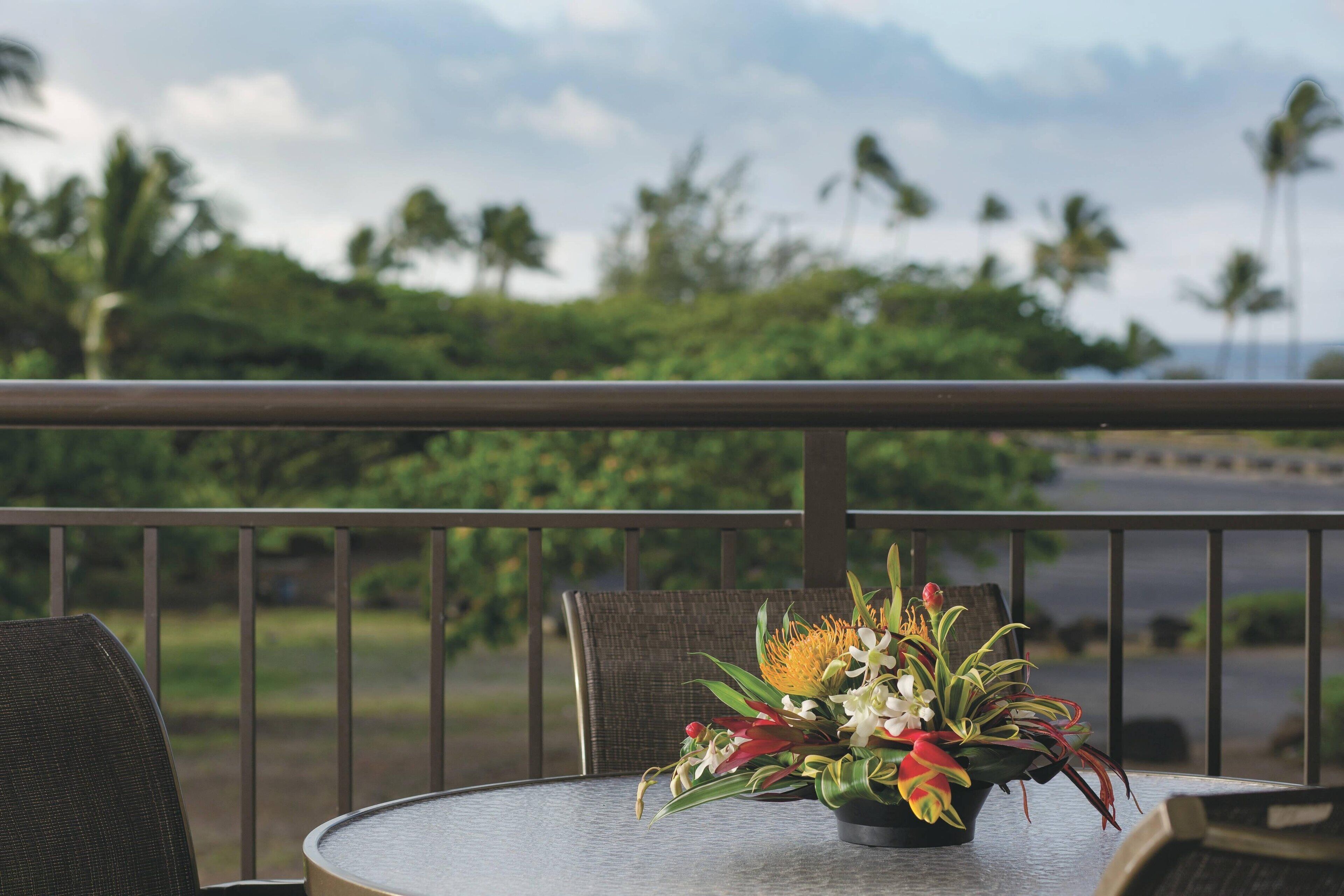 A round table with a floral arrangement on a balcony overlooking a scenic view with palm trees and a road.