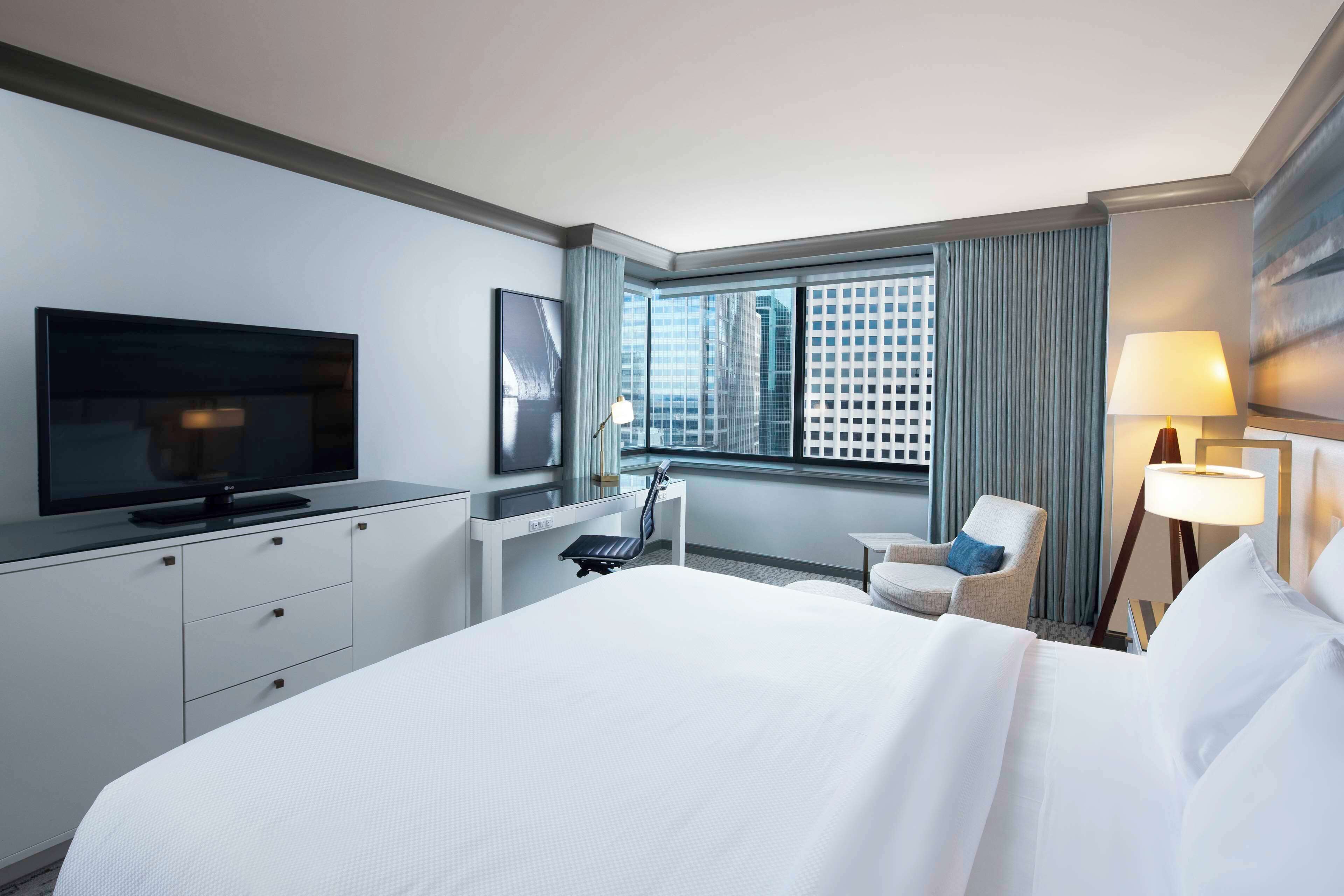 Quarto de hotel com cama grande, escrivaninha, televisão de tela plana, cadeira, janela com cortinas e abajur.