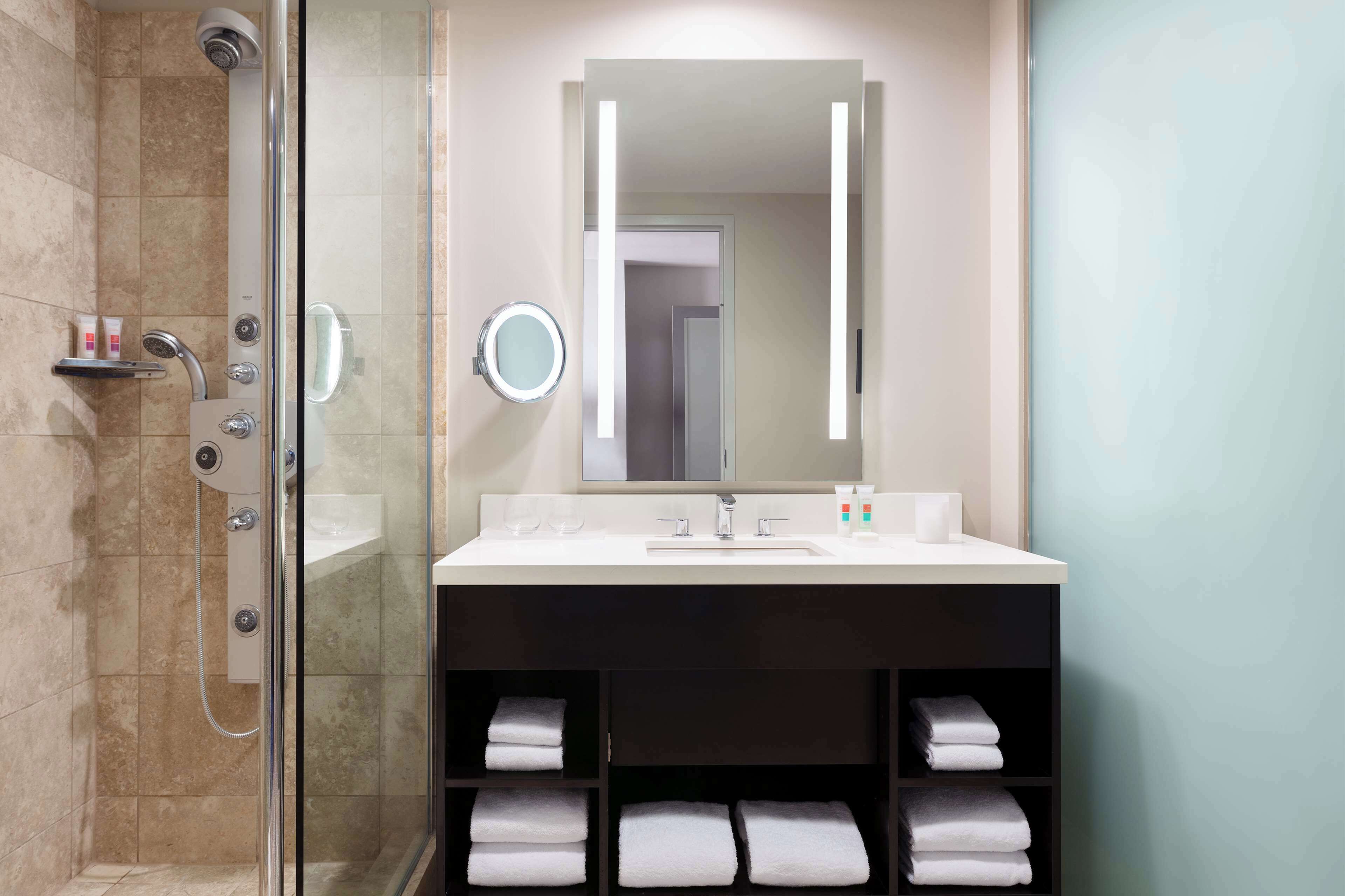 Un baño moderno con ducha de vidrio, un espejo grande y un tocador con lavamanos y espacio para toallas.