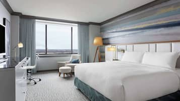 A hotel room with a large bed, a desk, a chair, a small round table, and a large window offering a city view.