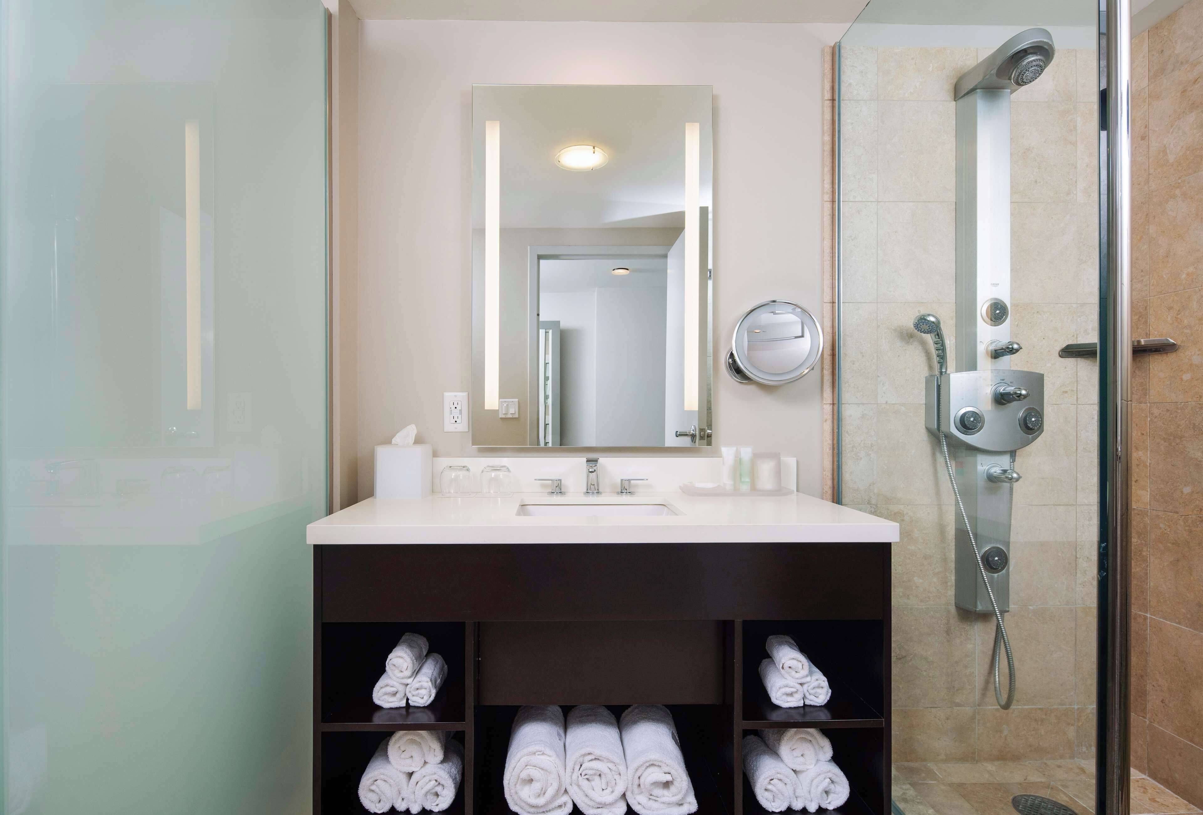 Une salle de bain moderne avec un grand miroir, un lavabo et une douche. Des serviettes sont soigneusement rangées sur des étagères situées sous le lavabo.