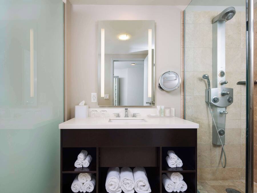 Une salle de bain moderne avec un grand miroir, un lavabo et une douche. Des serviettes sont soigneusement rangées sur des étagères situées sous le lavabo.