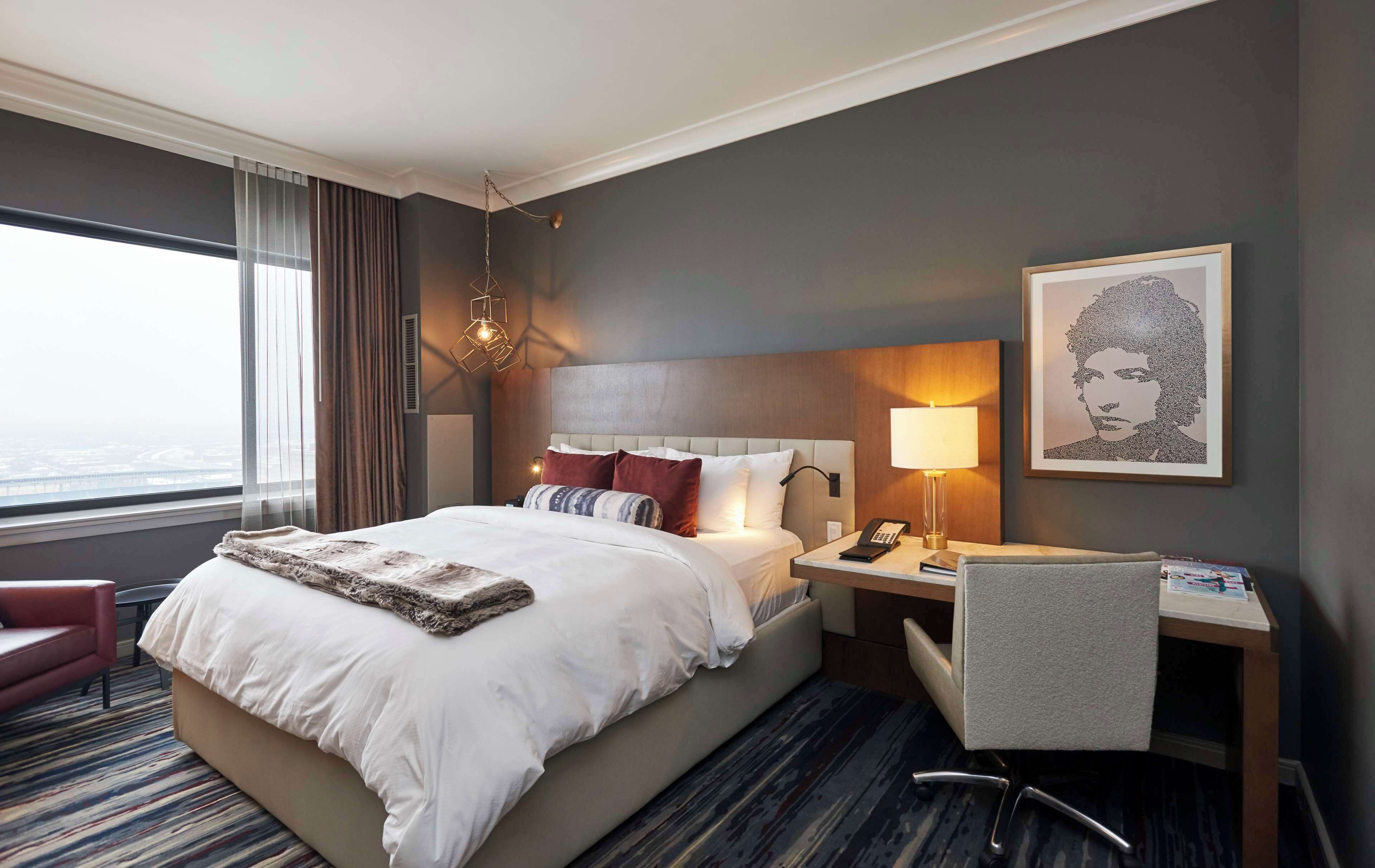 A hotel room with a large bed, a desk with a lamp, a chair, and a framed artwork on the wall.