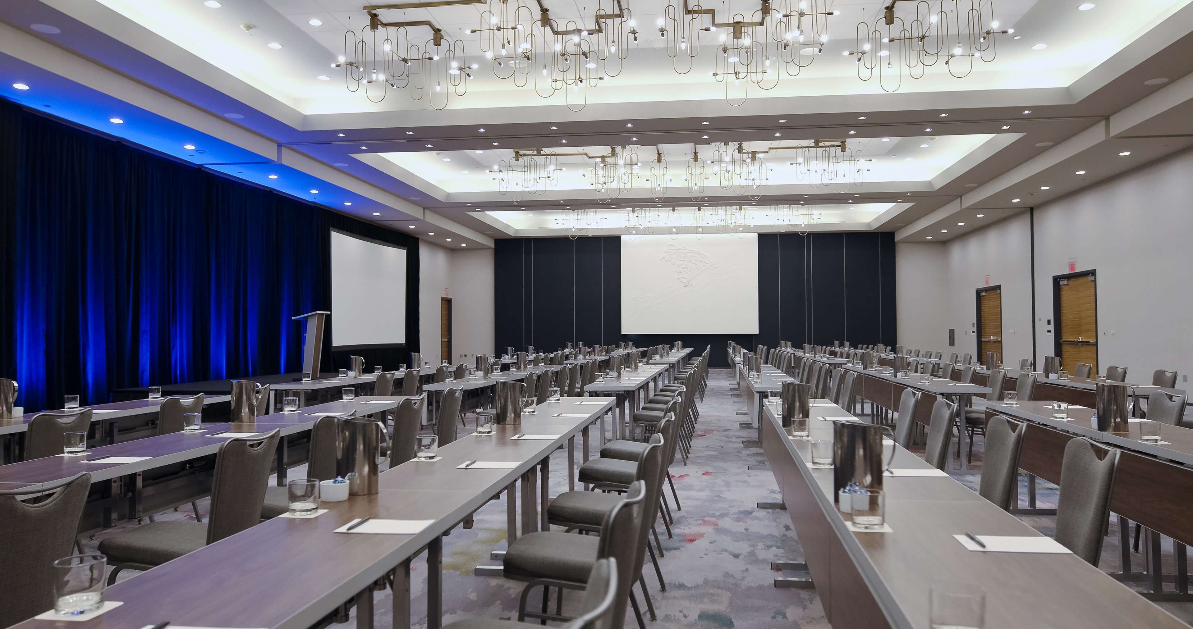 A conference room with long tables, chairs, and a large screen.
