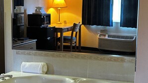 Un baño de hotel con una bañera grande, un espejo, un escritorio con silla, una lámpara y un equipo de aire acondicionado.