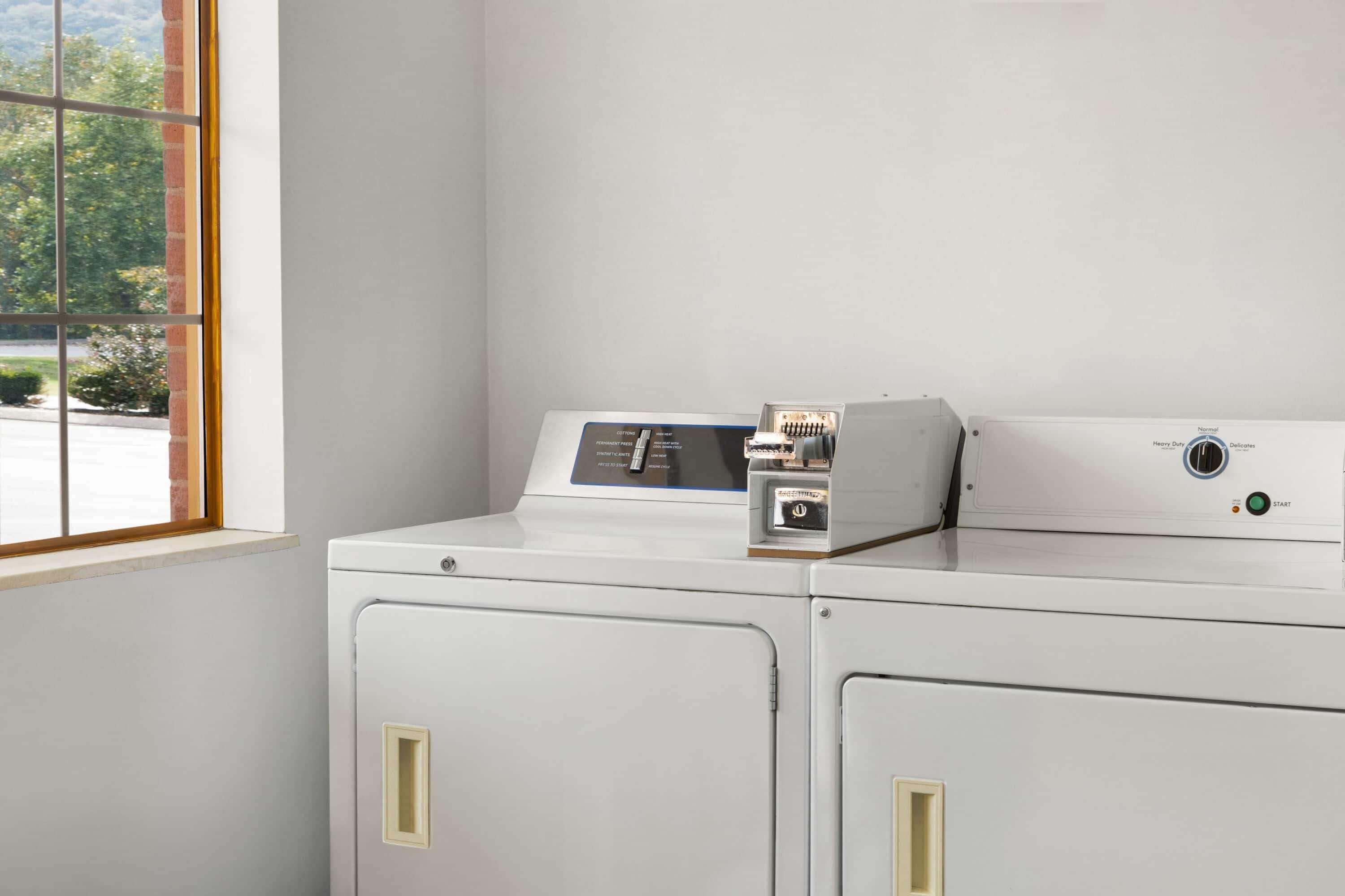 A laundry room with a front-loading washer and dryer, a control panel, and a window with a view of greenery outside.