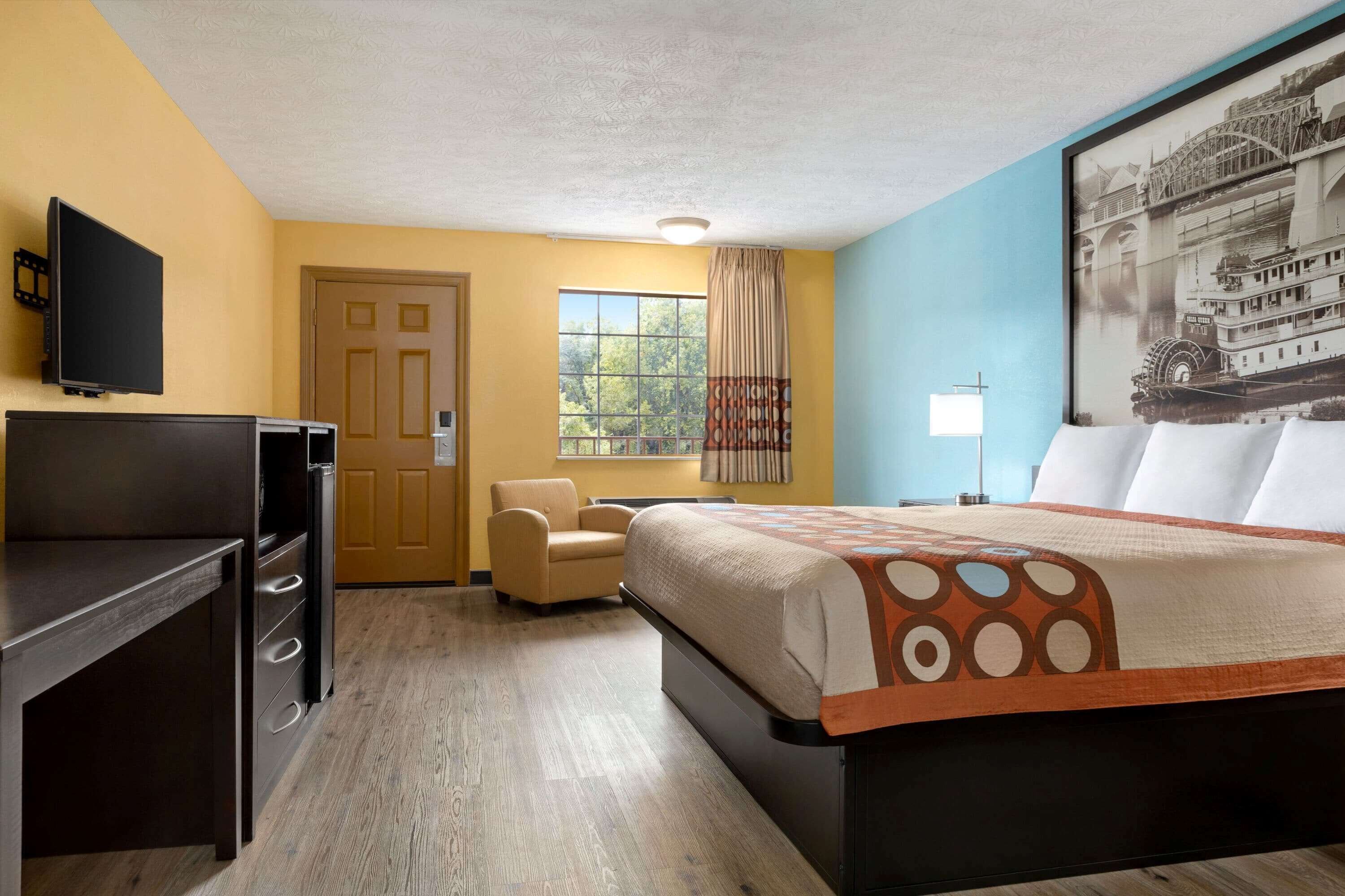 A hotel room with a bed, a desk, a chair, a window with curtains, and a framed picture on the wall.