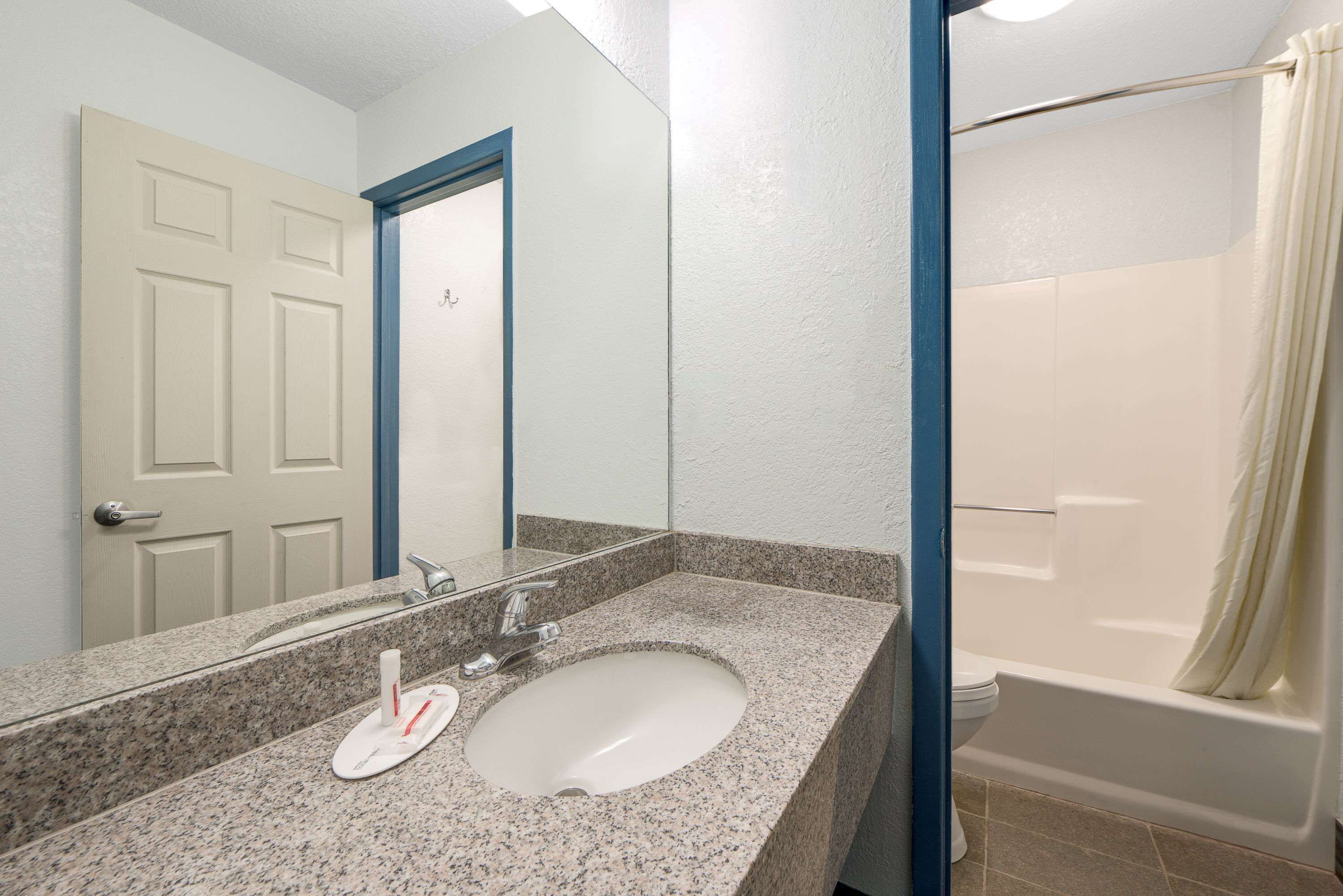 Baño con lavamanos de granito, espejo, inodoro blanco y bañera con cortina de ducha color beige.