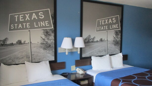 A hotel room with two beds, each with a Texas State Line sign above. There are two bedside lamps and a nightstand with a phone.