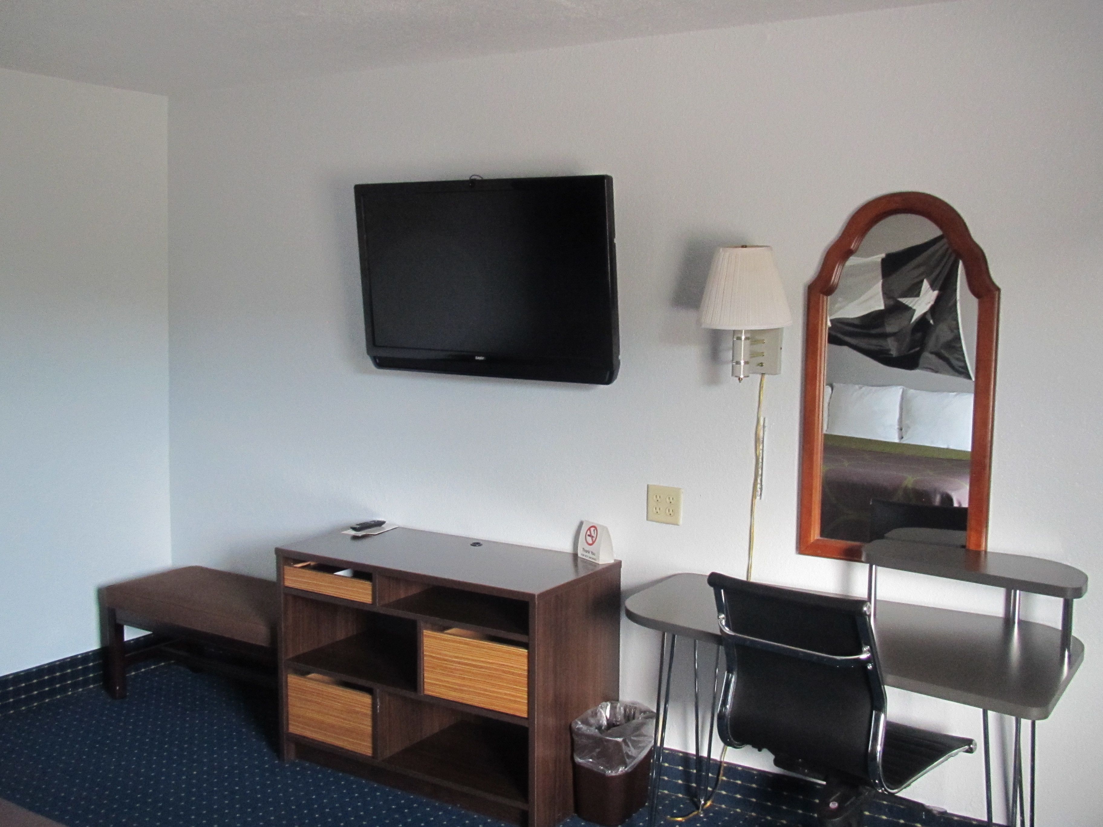A hotel room with a flat-screen TV mounted on the wall, a small table with a chair, a lamp, and a mirror with a star design.