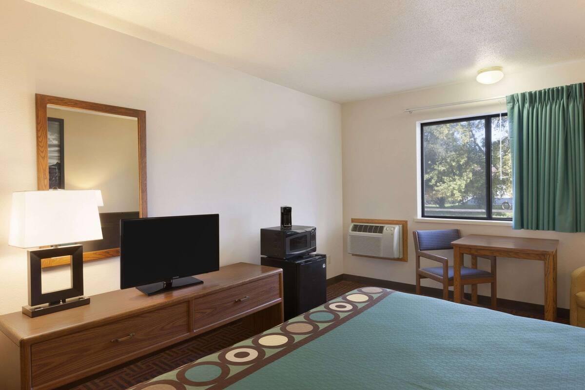 A hotel room with a bed, a TV, a desk, a chair, a lamp, a mirror, and a window with curtains.