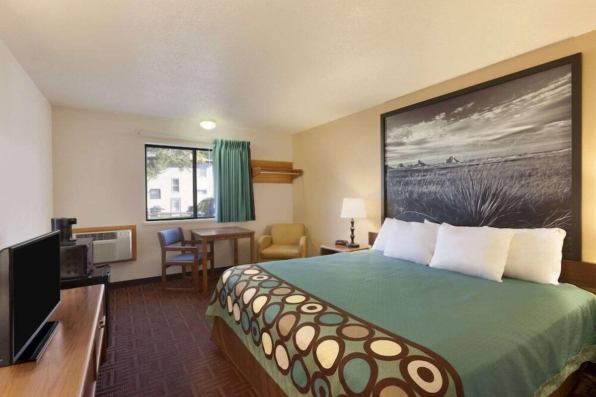 A hotel room with a bed, a desk with a computer, a chair, a window with curtains, and a painting on the wall.