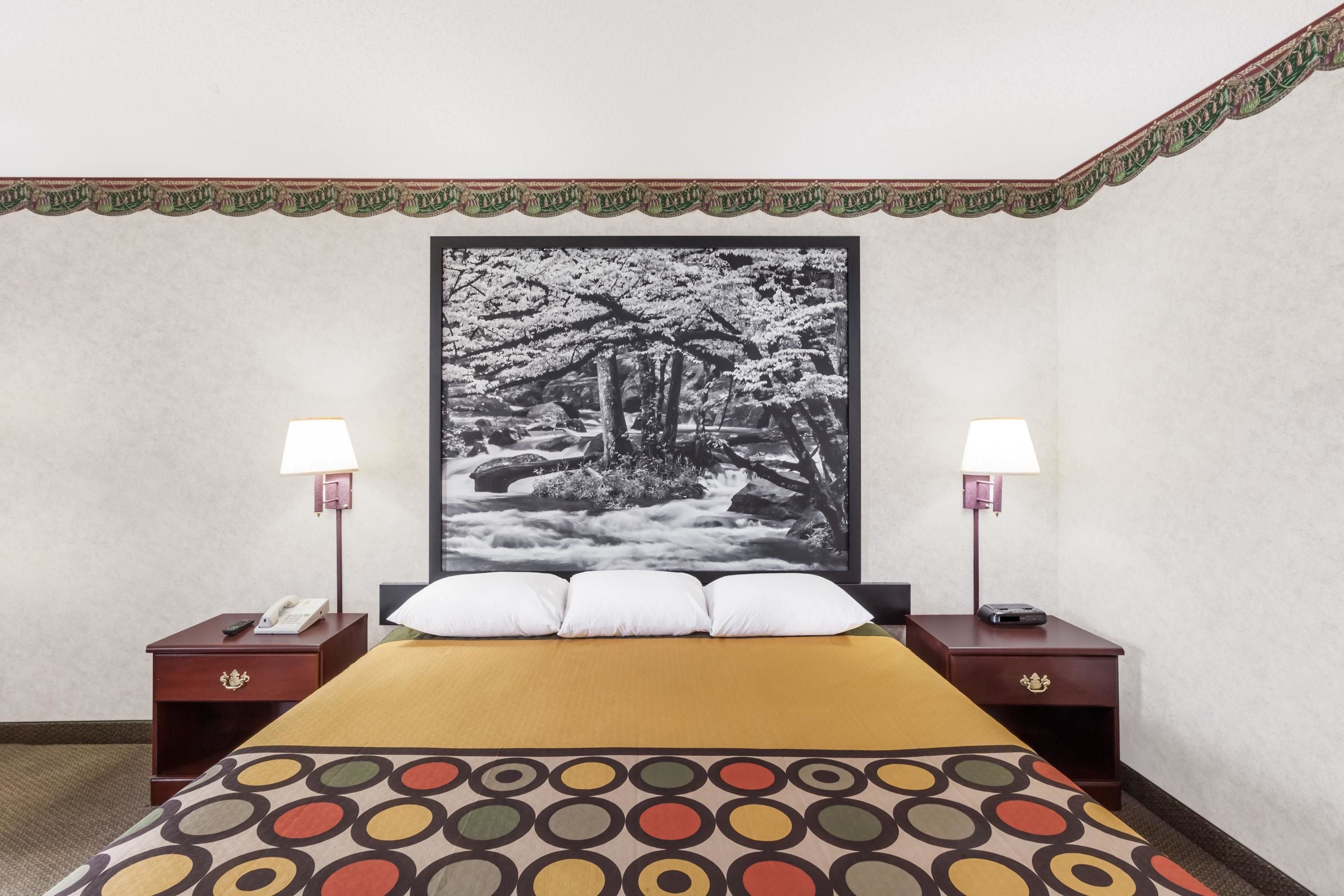 A hotel room with a bed, two bedside tables with lamps, a large framed picture on the wall, and a patterned bedspread.