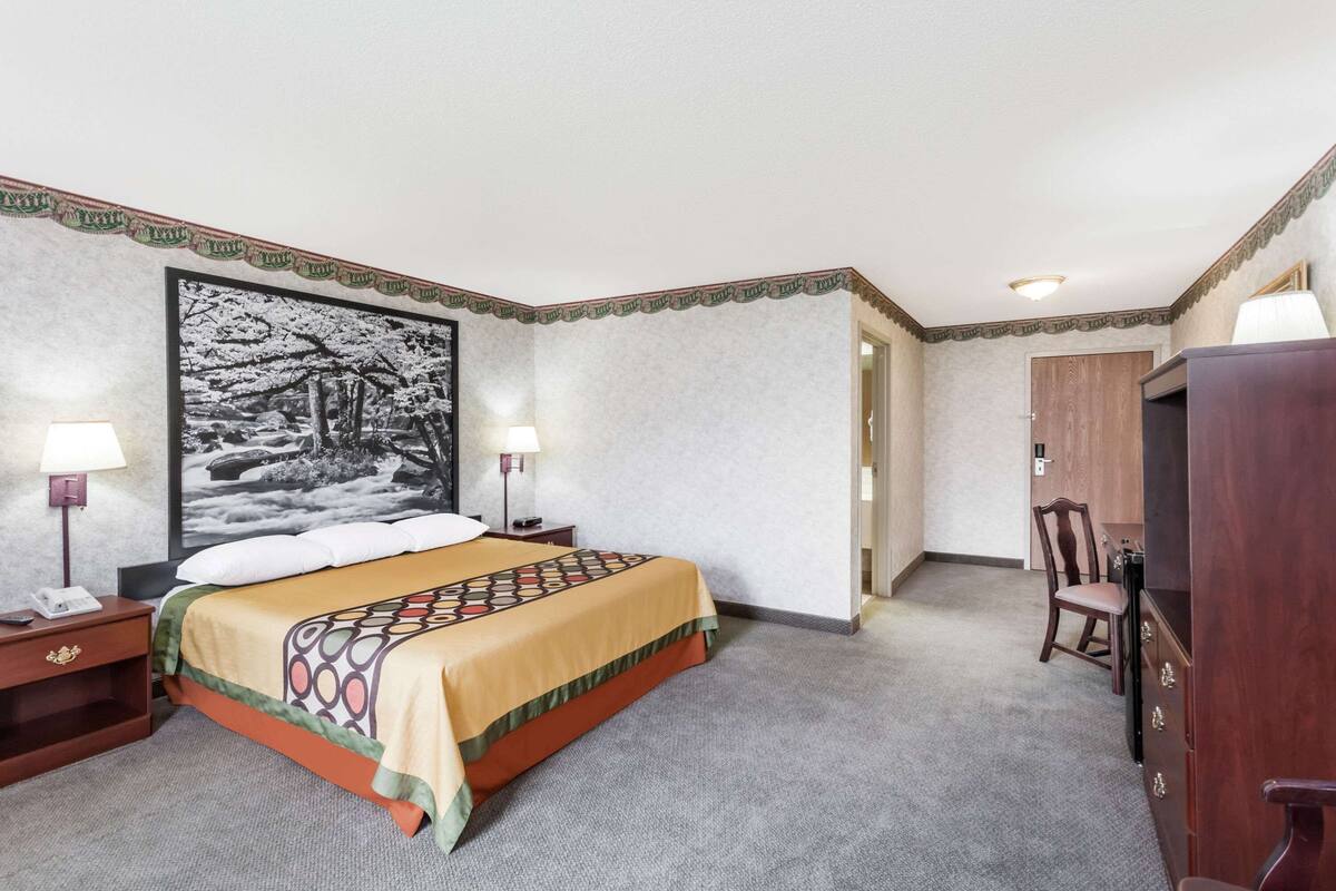 Une chambre d’hôtel avec un lit, deux tables de chevet avec des lampes, un grand tableau encadré au mur, une armoire en bois et une chaise.
