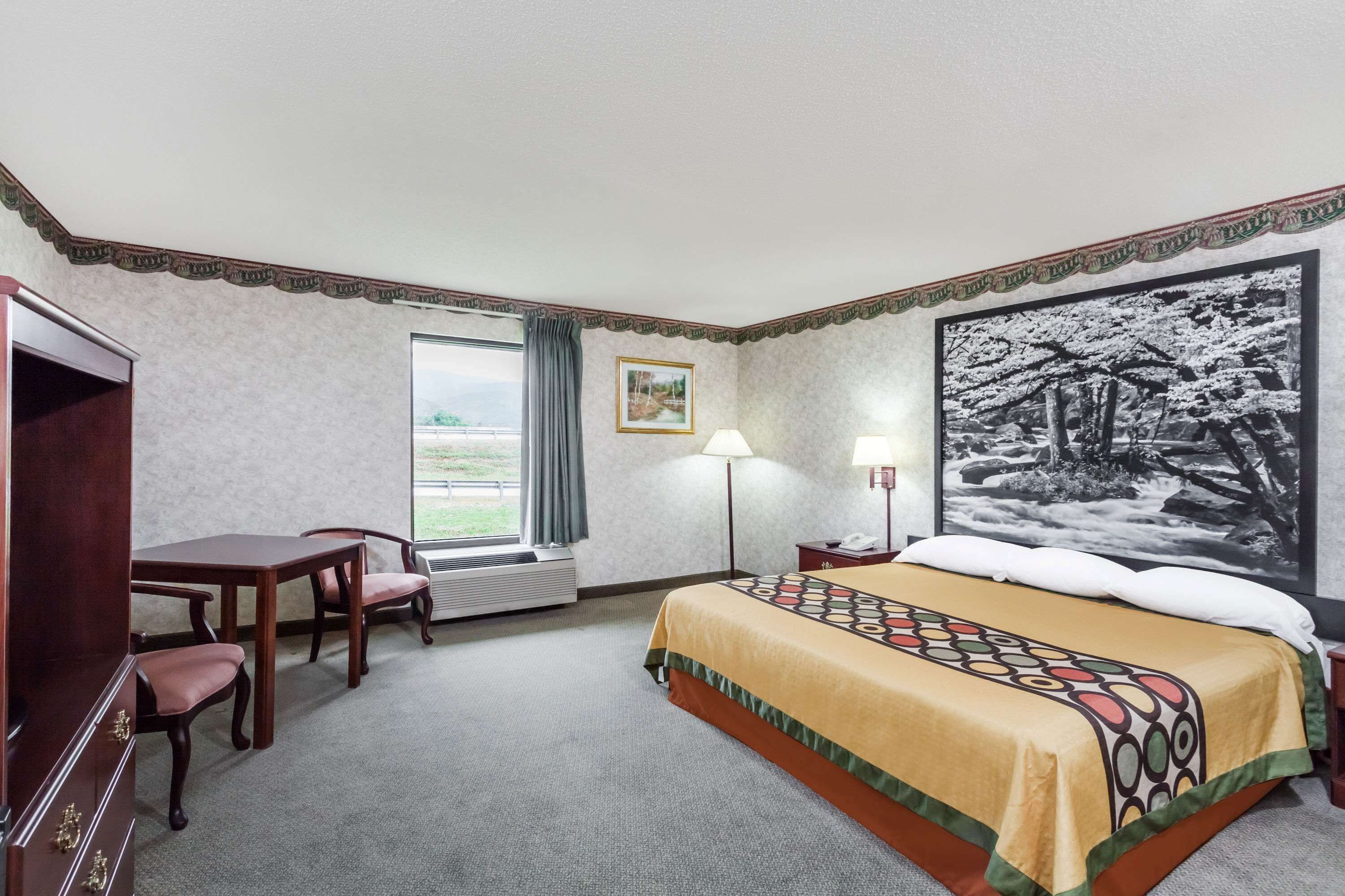 Una habitación de hotel con cama, escritorio, sillas, ventana con cortinas, lámpara y un cuadro en la pared.