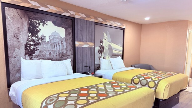 Een hotelkamer met twee bedden, elk met een gele sprei en witte kussens. Op de muur hangt een groot muurschildering van een historisch gebouw en een modern vliegtuig.
