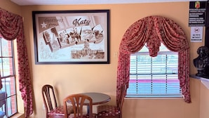 Een eethoek met een tafel en stoelen, een lijst met een foto aan de wand, behang met een patroon en een raam met zonwering.