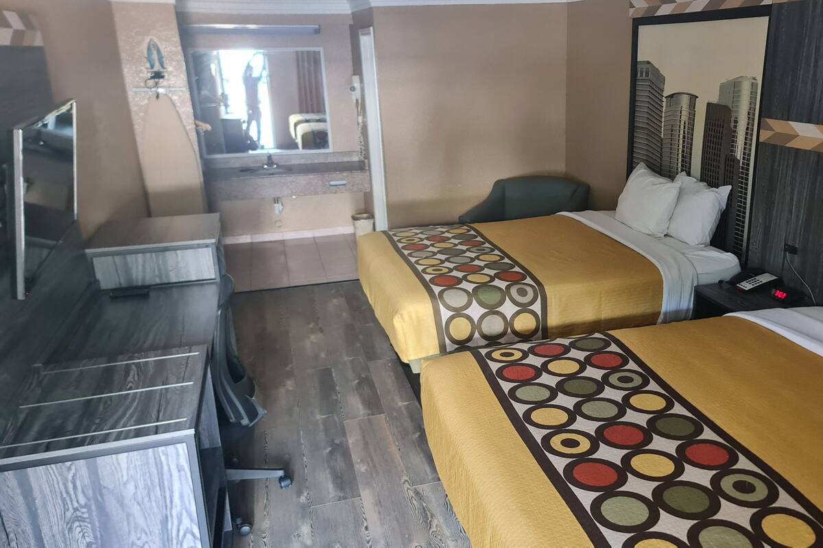 A hotel room with two beds, a desk, a chair, a mirror, and a view of buildings.