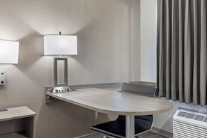 Habitación de hotel con una mesa redonda, una silla, una lámpara, un enchufe y una ventana con cortinas.