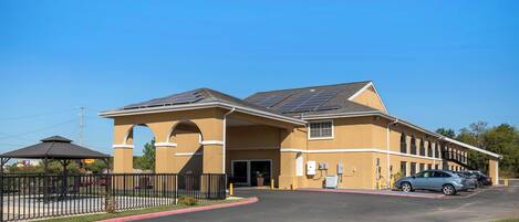 A building with a covered entrance, solar panels on the roof, and a parking area with cars.