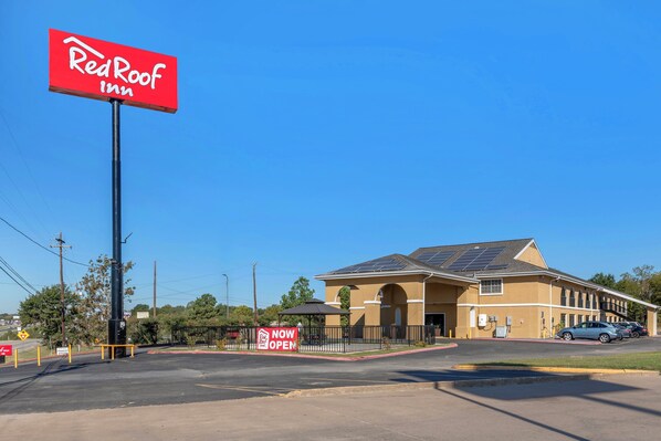 Un hotel Red Roof Inn bajo un cielo azul despejado, con paneles solares en el techo y un estacionamiento lleno de coches.