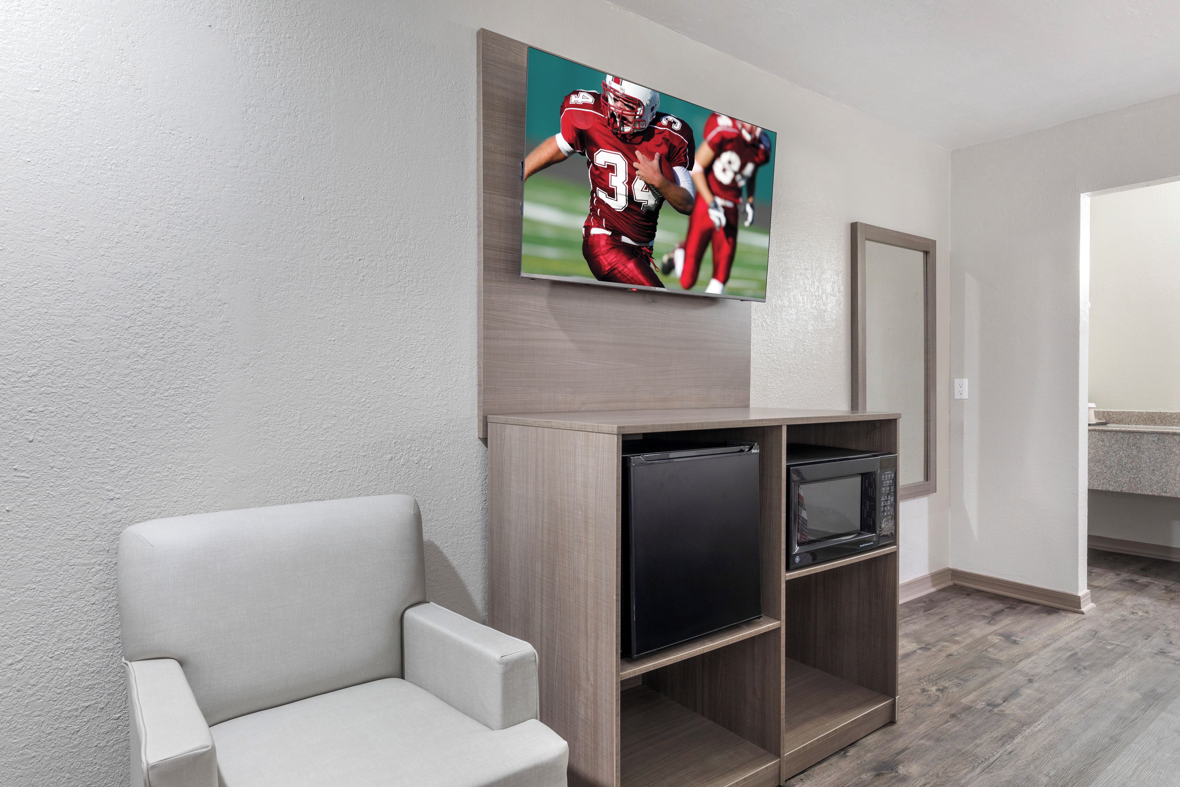 A hotel room with a flat-screen TV mounted on the wall, a small black refrigerator, and a grey armchair.