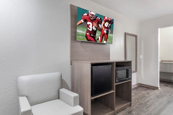 Habitación de hotel con televisor de pantalla plana en la pared, un refrigerador negro pequeño y un sillón gris.