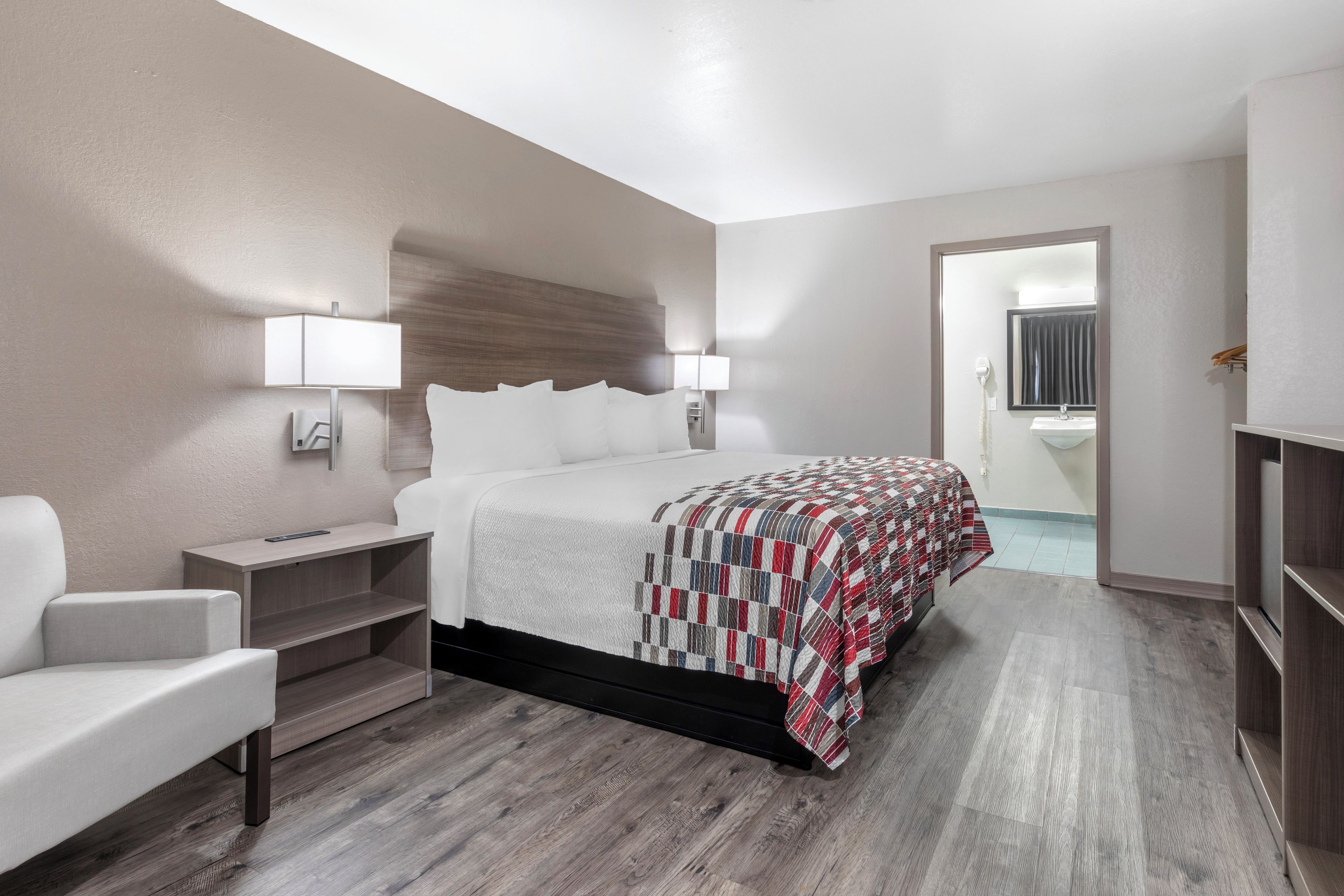 Una habitación de hotel con cama, mesitas de noche, una silla, armario y un baño visible a través de una puerta abierta.