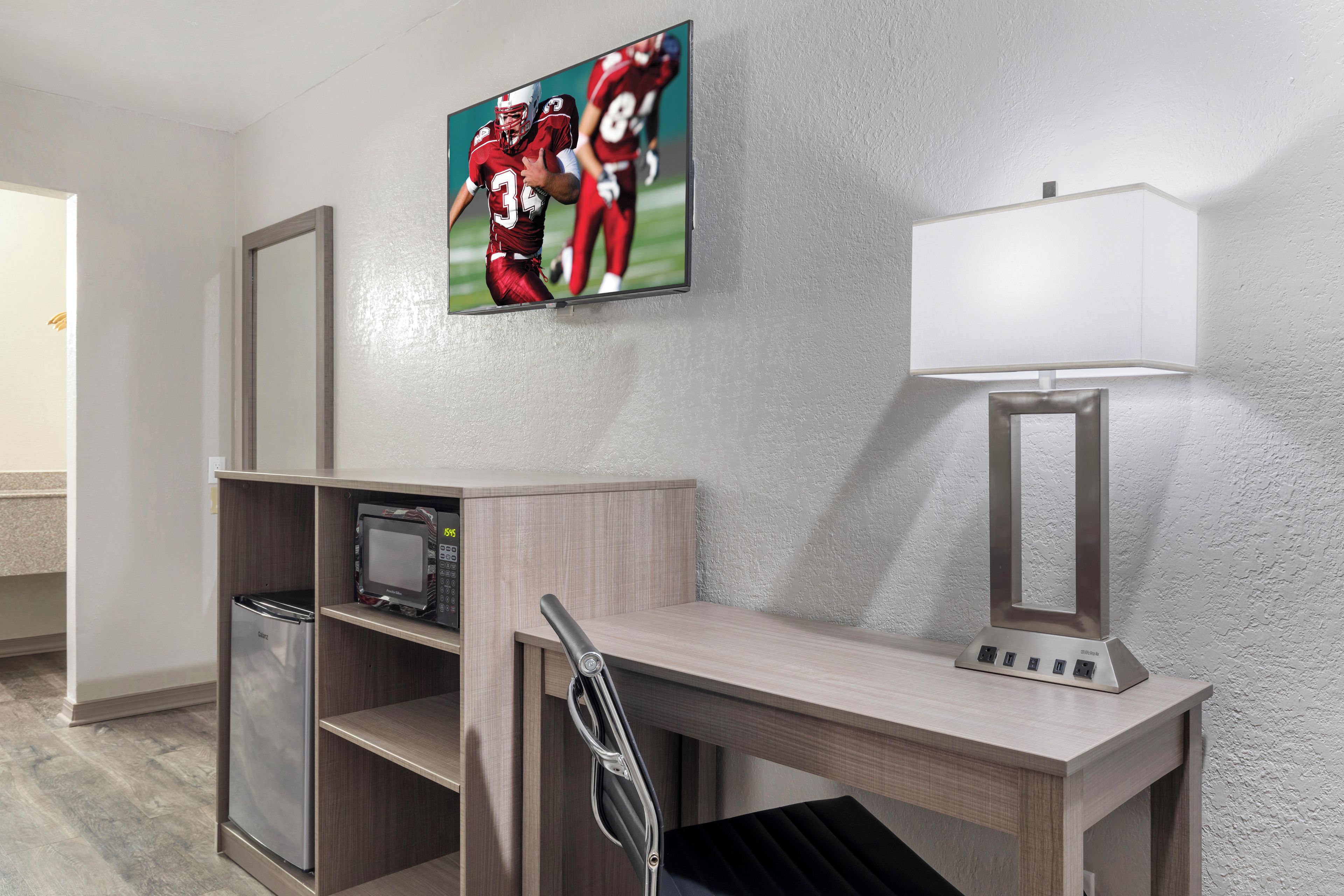 A hotel room with a desk, a microwave, a chair, a lamp, and a framed sports picture on the wall.