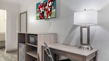 A hotel room with a desk, a microwave, a chair, a lamp, and a framed sports picture on the wall.